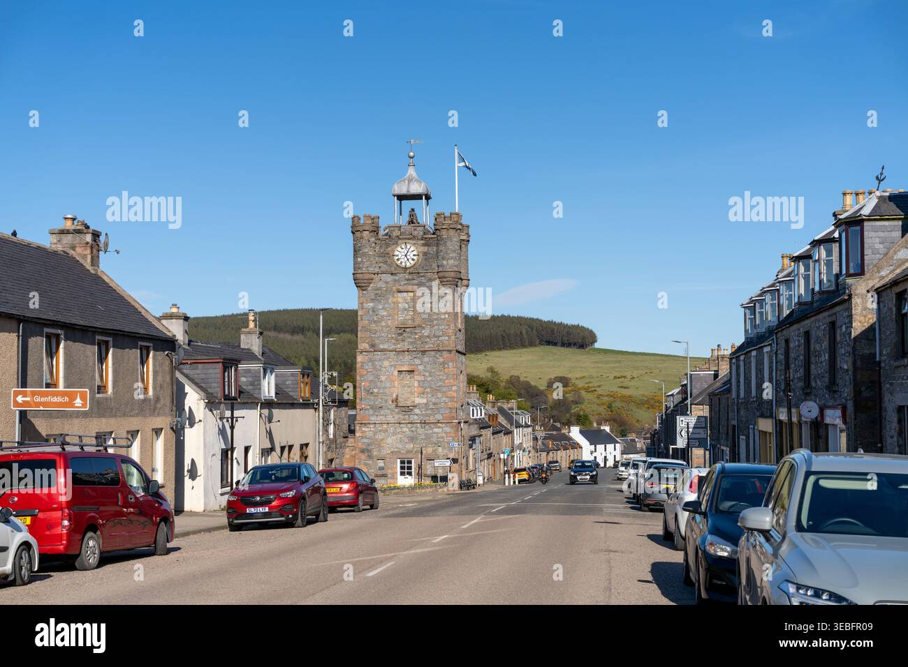 Ein klarer blauer Himmel hängt über Dufftown, Moray, Schottland, wo der historische Clock Tower fünf nach fünf zeigt. In einer belebten Straße unten gibt es Traditiona Stockfoto