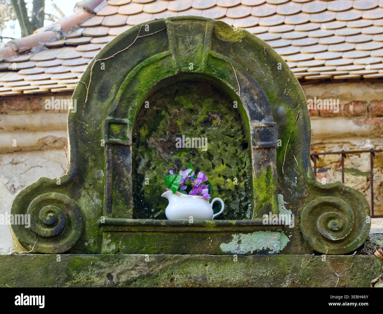 Kleiner moosbedeckter Steinschrein an einer Friedhofsmauer mit einem weißen Krug und Blumen als Hommage. Stockfoto
