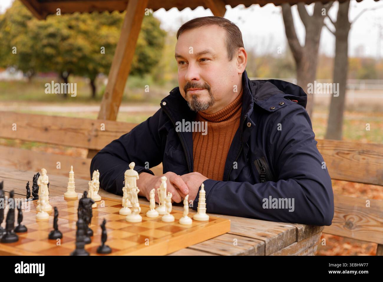 Konzentrierter reifer Mann, der an einem Holztisch in einem Herbstpark sitzt und mit dekorativen Stücken ein Schachspiel spielt. Konzept der Strategie, des Hobbys und des Geistes Stockfoto