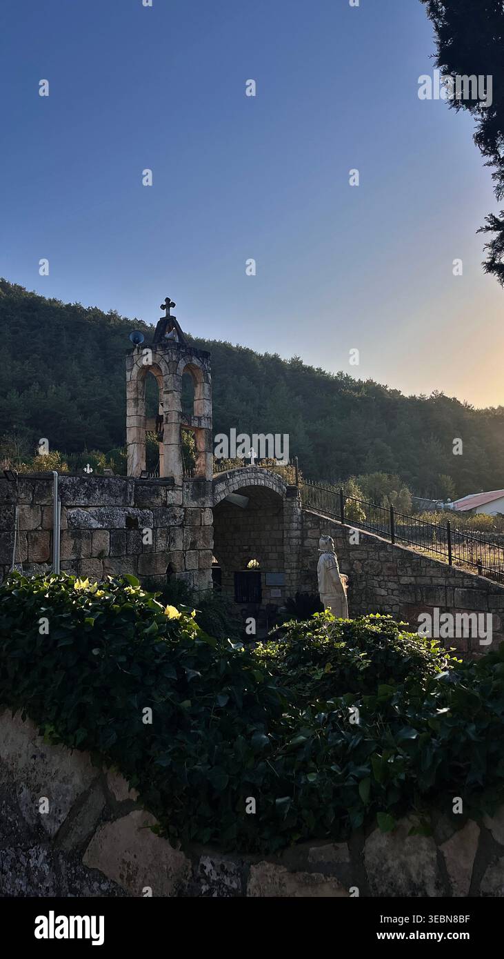 Ruinen der Kirche Saint Challita bei Sonnenuntergang, Kobayat, Nordlibanon. Steinglockenturm, bogengewölbtes Tor und mit Efeu bedeckte Mauern. - Smartphone-aufgenommenes Stockfoto