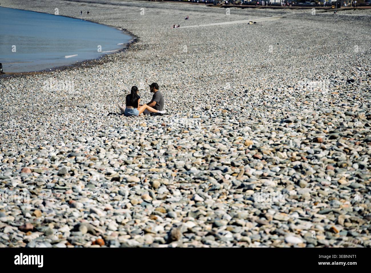 Unter einem klaren blauen Himmel säumen glatte Kieselsteine eine ruhige Bucht von Llandudno. Ein Paar sitzt wie Kieselsteine am Strand. Stockfoto