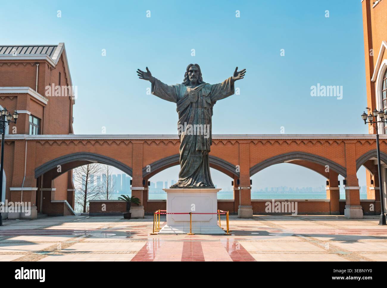 Jesus Christus Statue mit ausgebreiteten Armen steht auf einem sonnigen platz vor einer Backsteinkirche mit Blick auf das Wasser und eine ferne Stadt Stockfoto