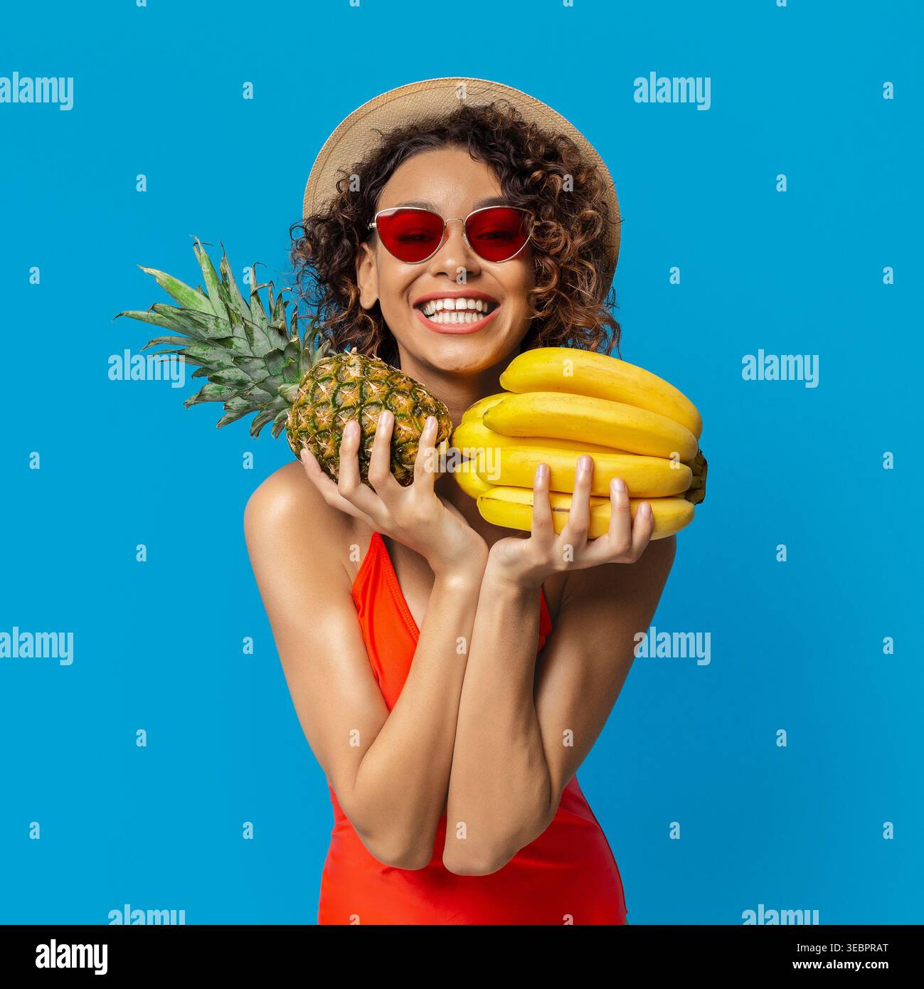 Eine junge Frau mit lockigem Haar und roter Sonnenbrille lächelt weit und hält Ananas und Bananen in der Hand Stockfoto