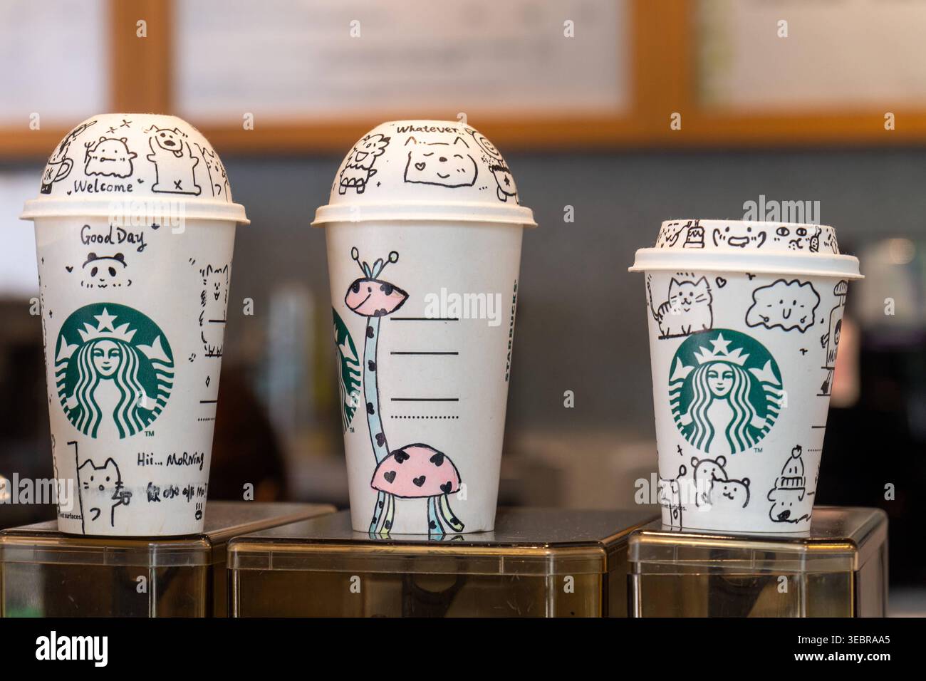 Hongkong, China - 25. April 2026: Drei Coffee-to-go-Tassen mit dem Starbucks Logo und kundenspezifischen Illustrationen stehen auf einer Theke in einem cafÃ. Die Einwegbecher, die in verschiedenen Größen erhältlich sind, sind für Heißgetränke wie Kaffee oder Tee bestimmt *** drei Coffee-to-Go-Becher mit Starbucks-Logo und individuelle Illustrationen stehen auf einer Theke in einem CafÃ. Die verschiedenen großen Einwegbecher sind für Heißgetränke wie Kaffee oder Tee vorgesehen Stockfoto