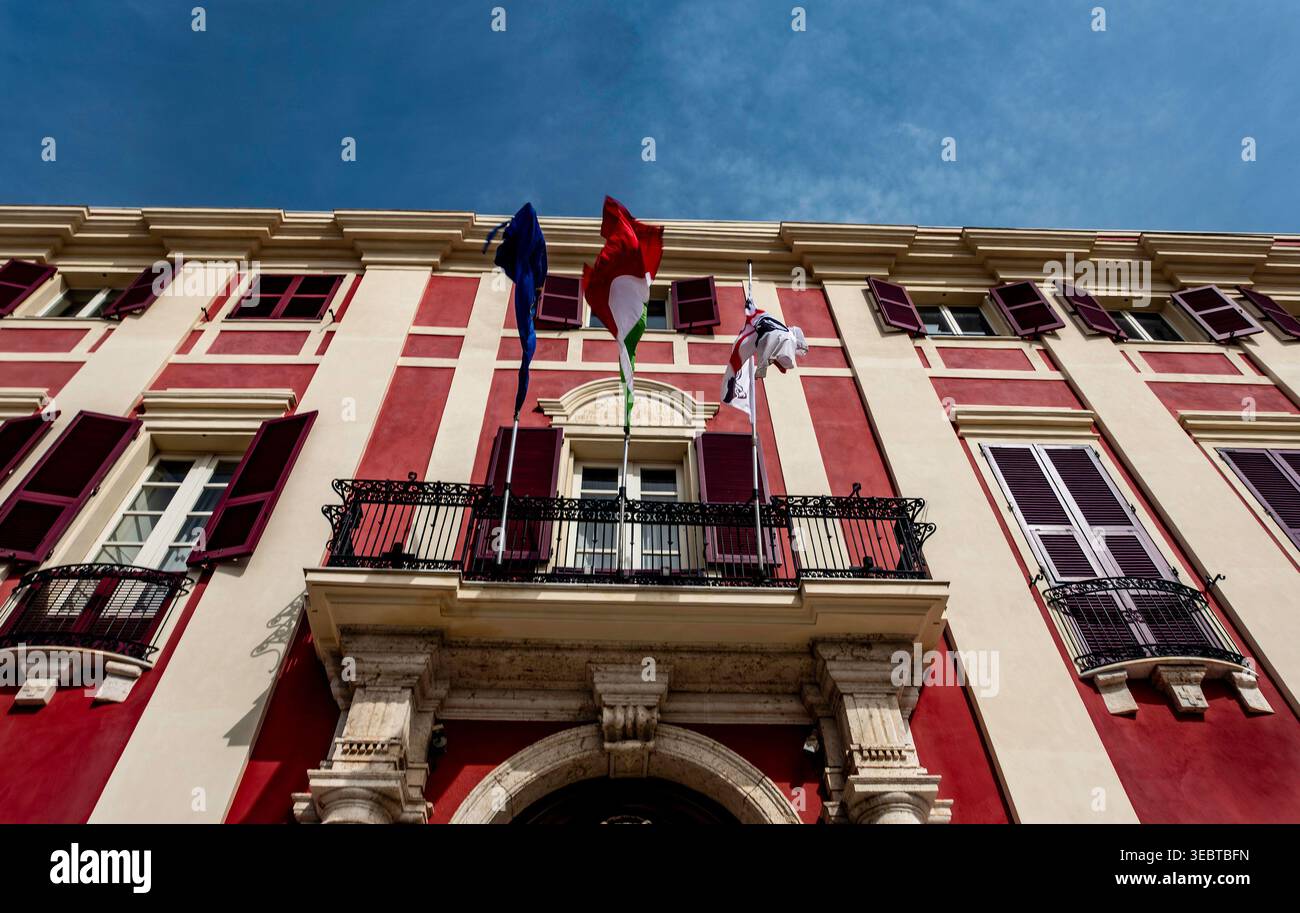 Fahnen fliegen über einen Balkon im Palazzo Regio in Cagliari. Die historische Fassade hat rote Paneele, weiße Zierleisten und dunkle Fensterläden unter blauem Himmel. Stockfoto