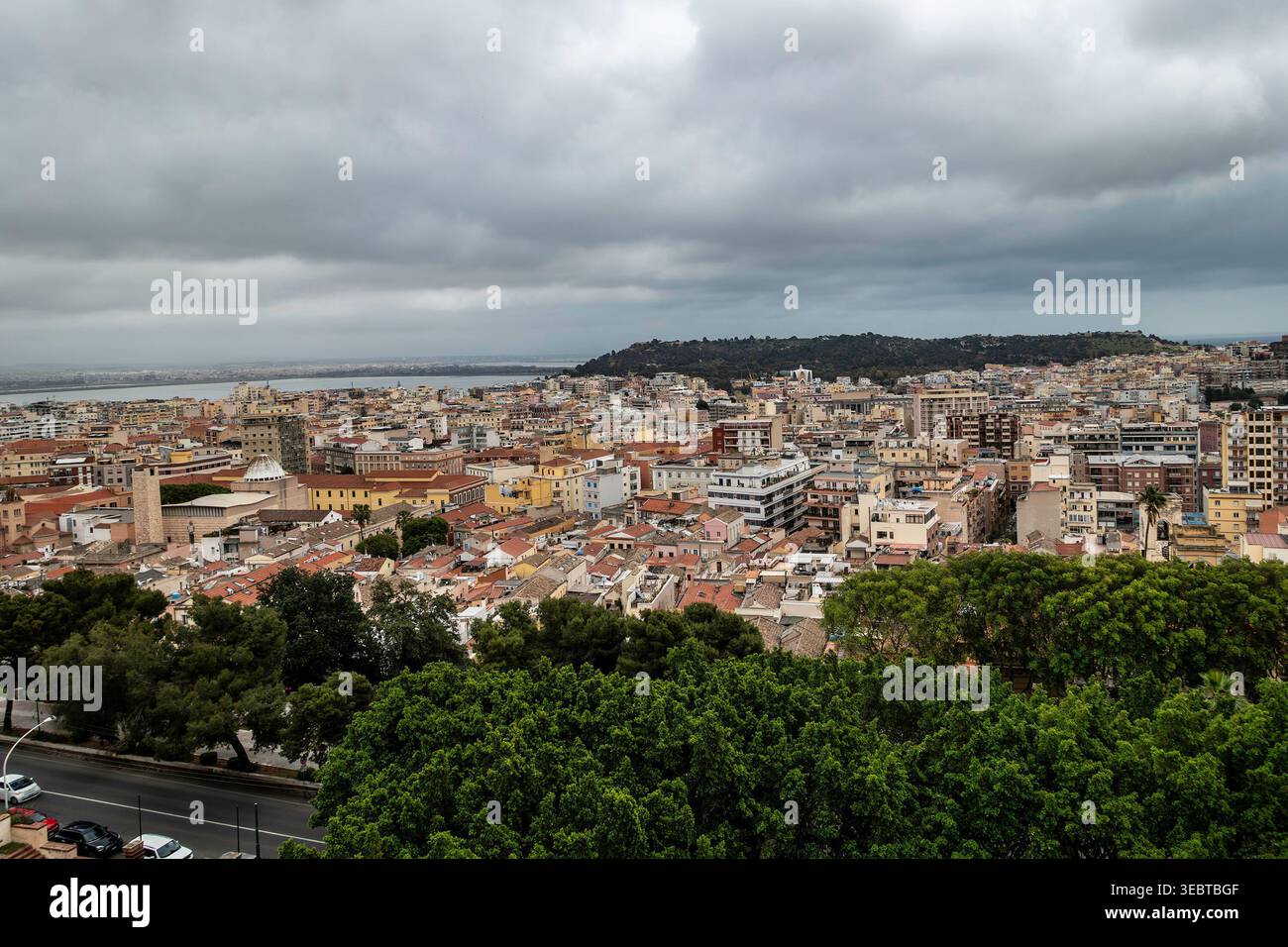 : Ein erhöhter Blick auf Cagliaris urbane Landschaft unter schweren grauen Wolken. Dichte Häuser, grüne Baumkronen und das ferne Meer bestimmen die Skyline. Stockfoto