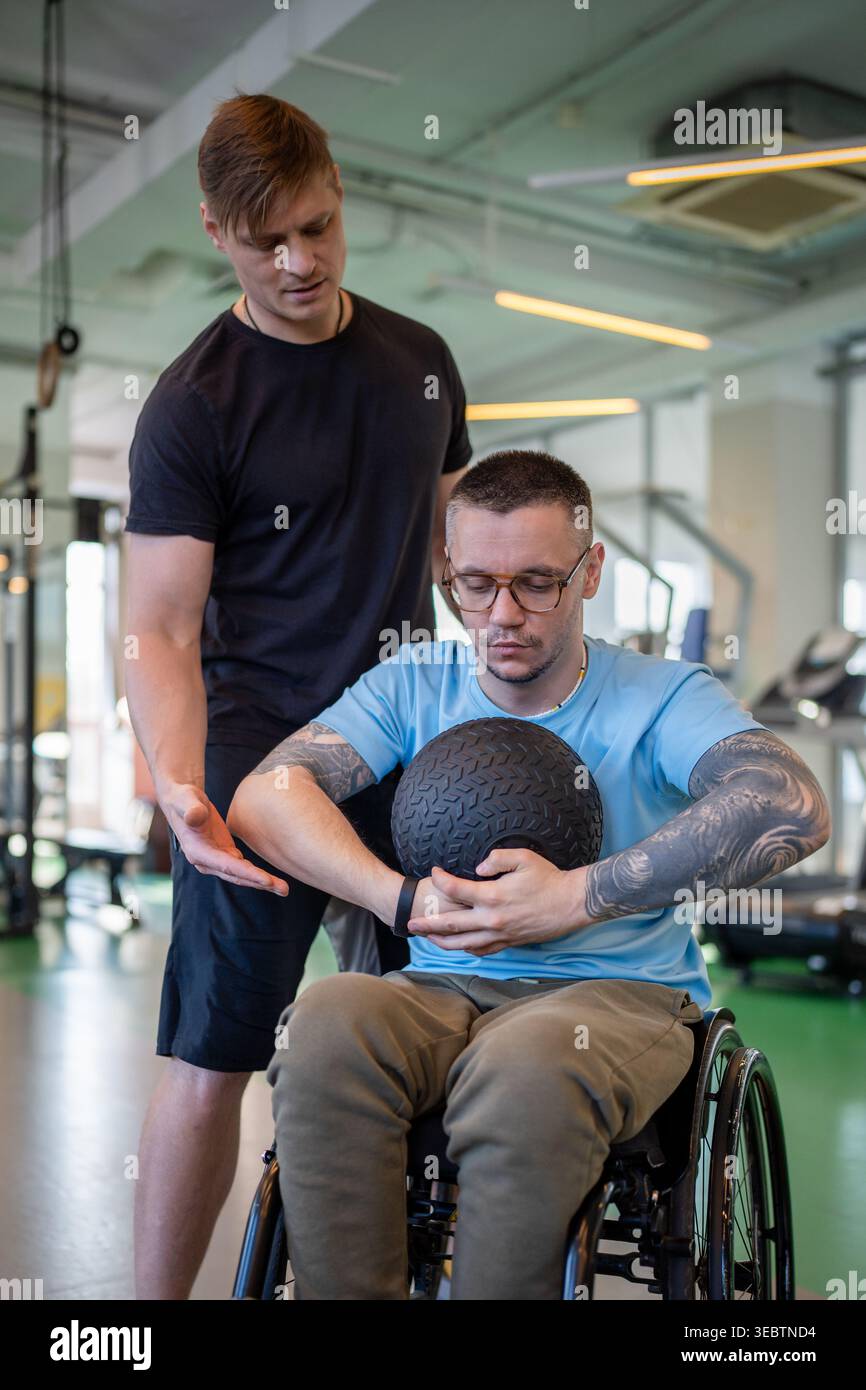 Personal Coach unterstützt behinderten Mann im Rollstuhl beim Krafttraining mit Medizinball im Fitnessstudio. Stockfoto