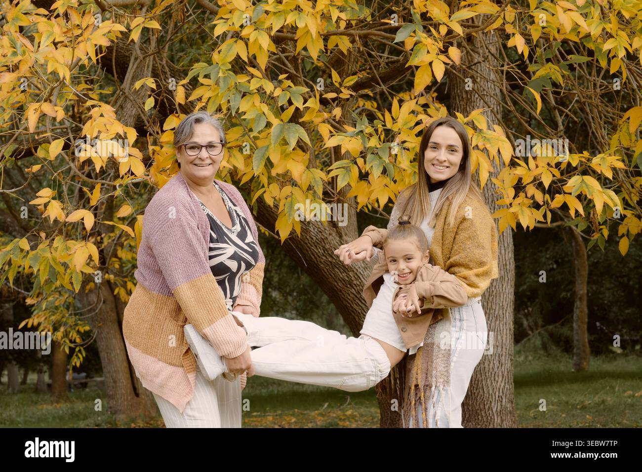 Großmutter, Mutter und Tochter spielen zusammen in einem Herbstpark, heben das Kind waagerecht unter goldenen Bäumen an und fangen eine fröhliche und fröhliche Stimmung ein Stockfoto