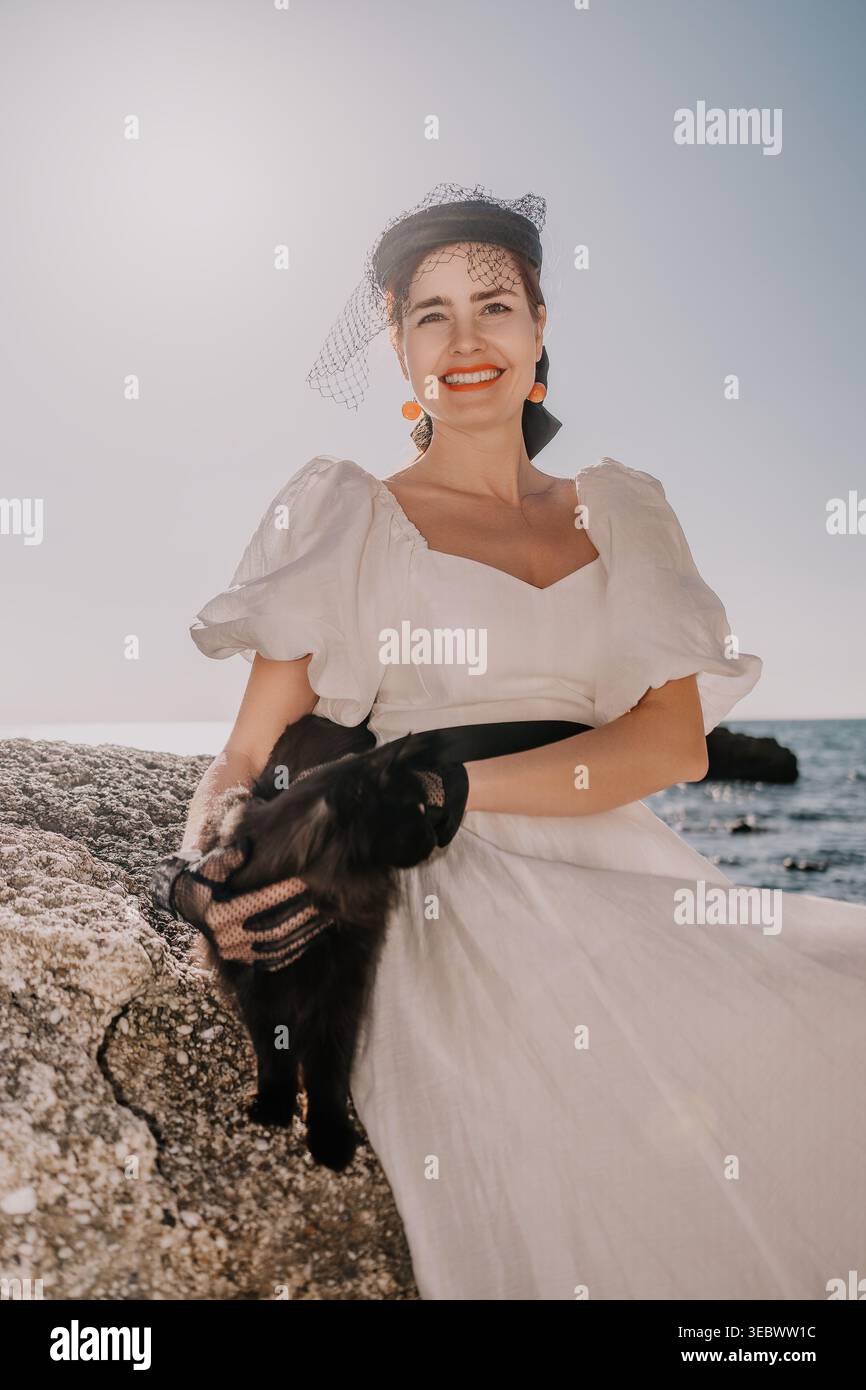 Frau Katze Küste fröhliche junge Dame in elegantem weißem Kleid und Hut mit einer schwarzen Katze an der felsigen Küste bei Sonnenuntergang Stockfoto