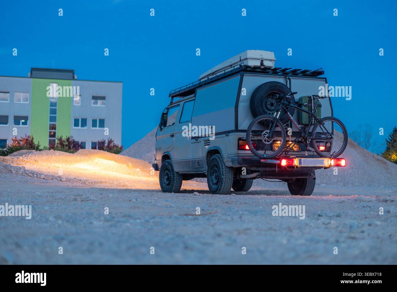 Ein robuster Geländewagen, der in der Abenddämmerung auf Schotter geparkt ist und mit einem Dachträger, Mountainbike und Reserverad ausgestattet ist. Ein modernes Gebäude mit grünen Akzenten Stockfoto