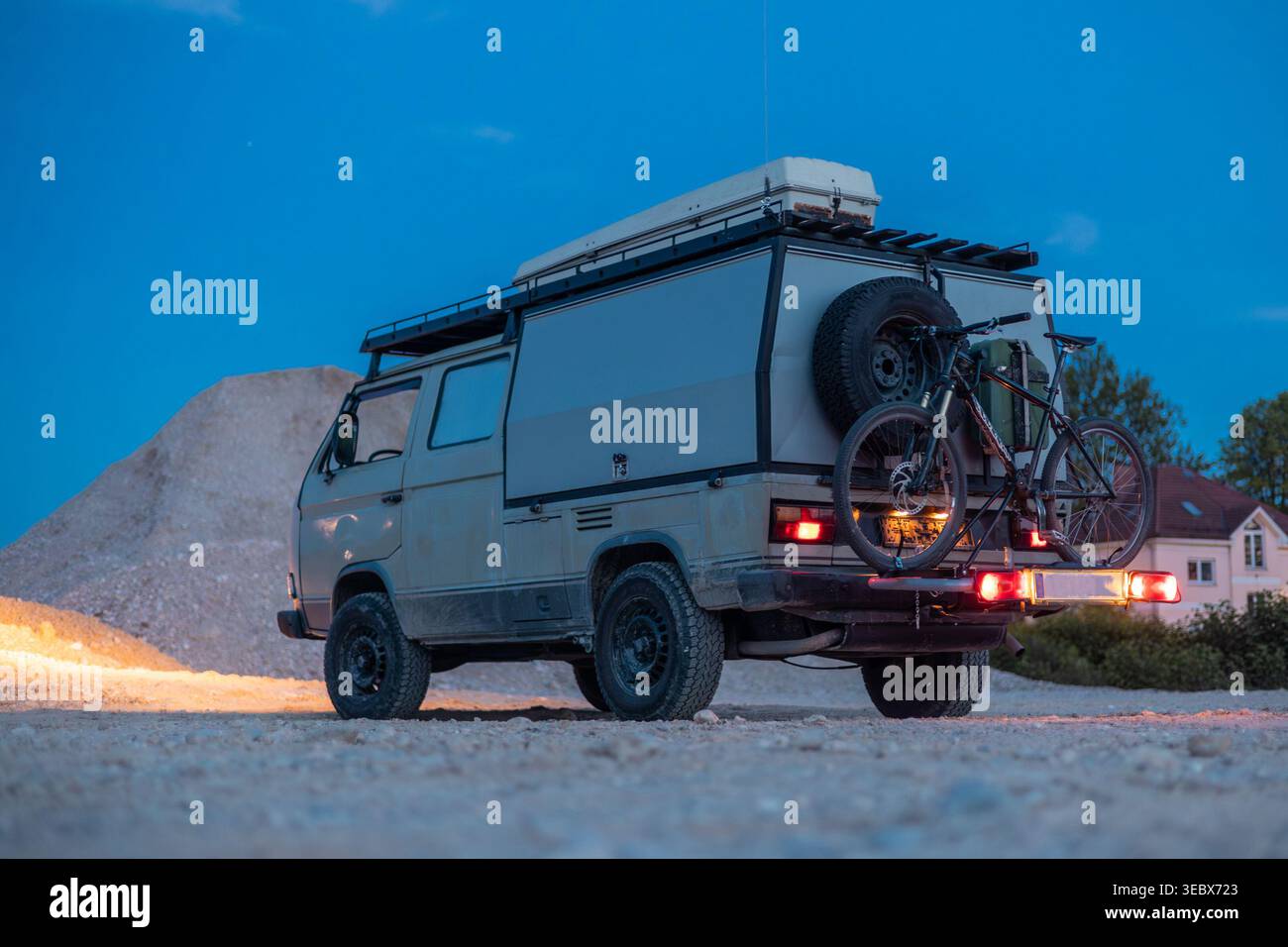 Ein robuster Geländewagen, der in der Abenddämmerung auf Schotter geparkt ist und mit einem Dachträger, Mountainbike und Reserverad ausgestattet ist. Ein modernes Gebäude mit grünen Akzenten Stockfoto