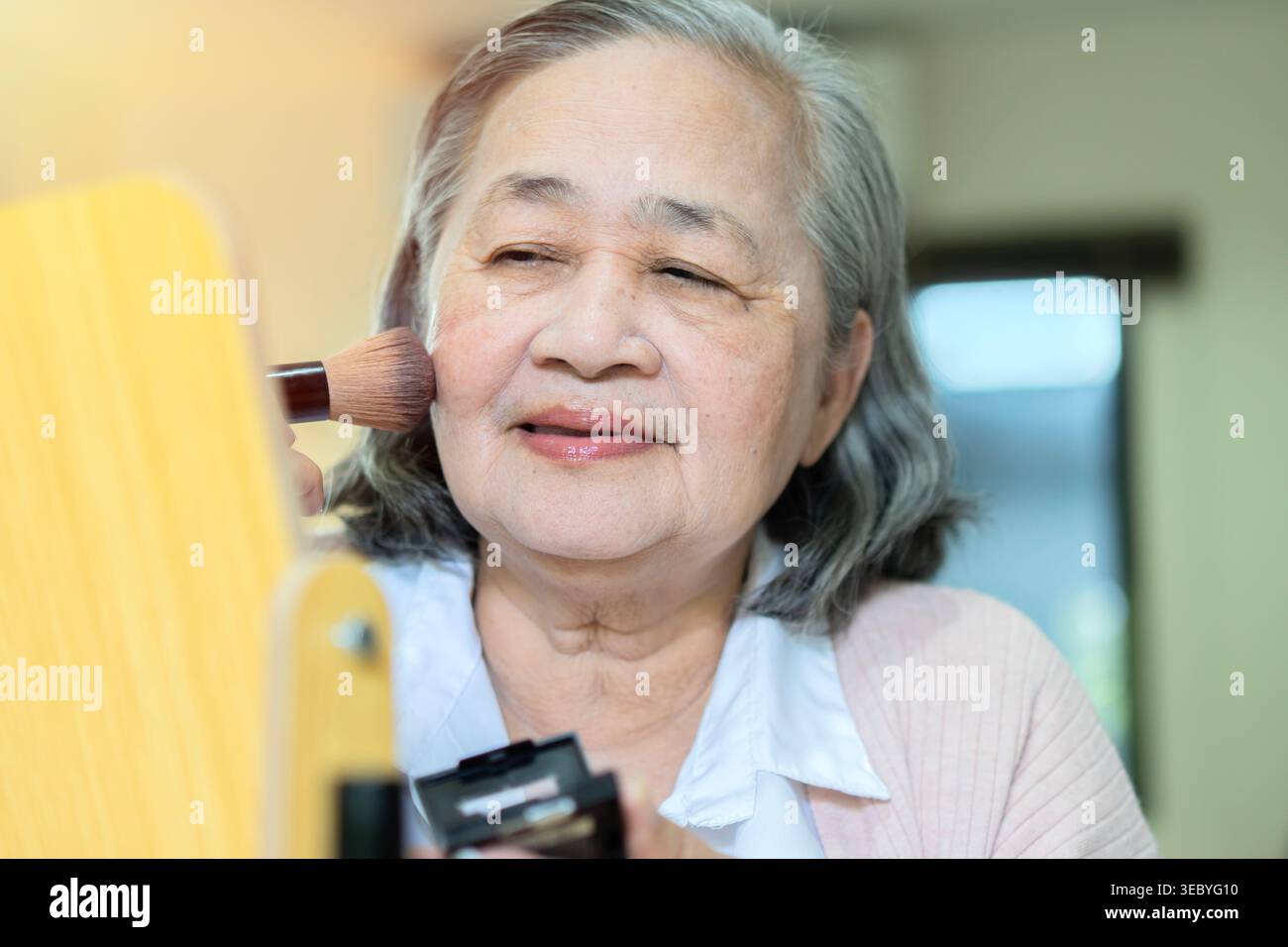 Eine schöne ältere asiatische Frau sitzt zu Hause und macht ihre Make-up-Routine am Morgen. Eine ältere Frau, die ihr Gesicht rötet Stockfoto