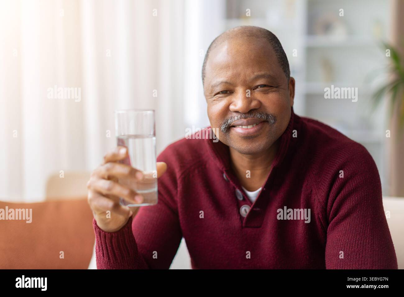 Lächelnder reifer schwarzer Mann, der ein Glas Wasser zu Hause hält Stockfoto