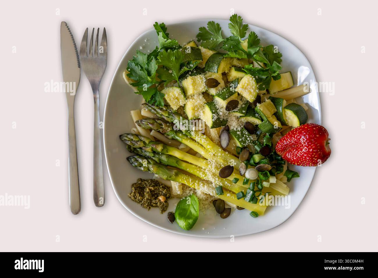 Blick von oben auf einen großzügigen Teller mit Pasta, grünem Spargel, Zucchini, Basilikum, Petersilie, Pesto, Parmesankäse, Kürbiskerne und eine Erdbeere, ein Kni Stockfoto