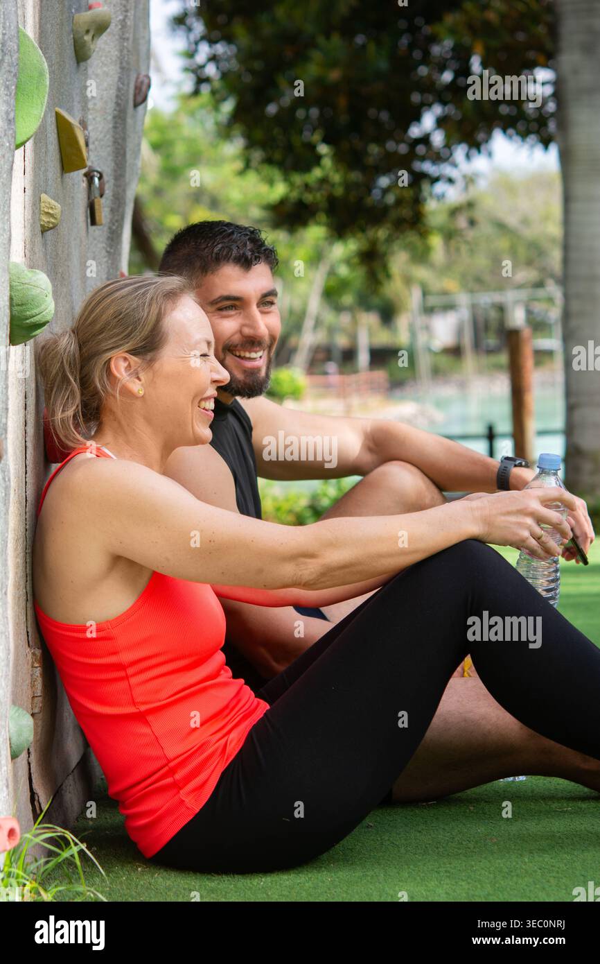 Glückliches, sportliches Paar, das nach dem Training im Freien sitzt und sich entspannt. Lächelnder Mann und Frau, die in der Nähe der Kletterwand ruhen, Wasser trinken und aktiv genießen Stockfoto