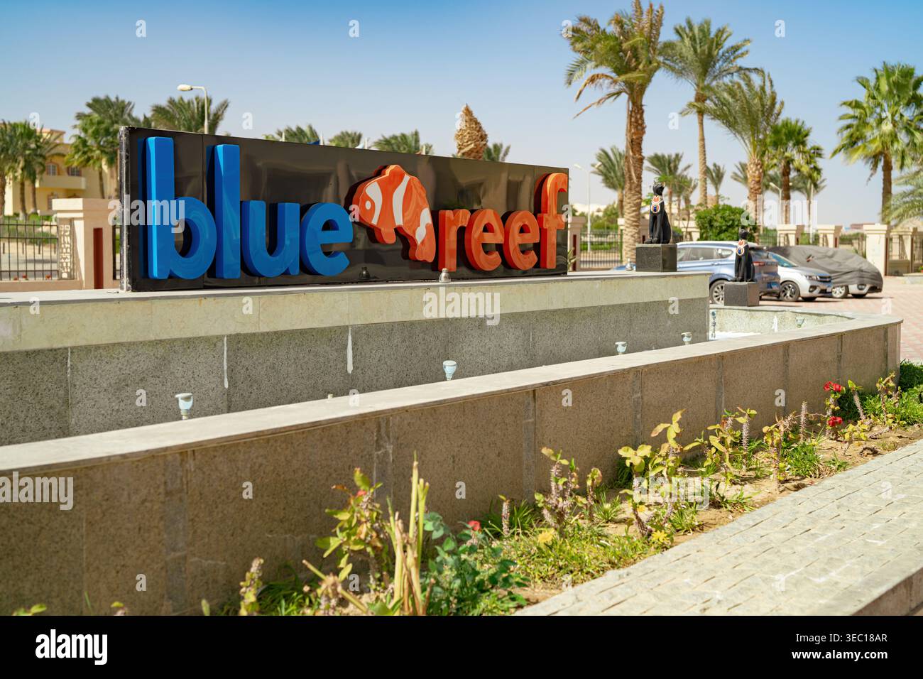 Marsa alam, ägypten 21. april 2026: Blaues Riffresort-Eingangsschild mit Clownfish-Logo, Palmen und Brunnen unter sonnigem rotem Meereshimmel in marsa alam, z. B. Stockfoto
