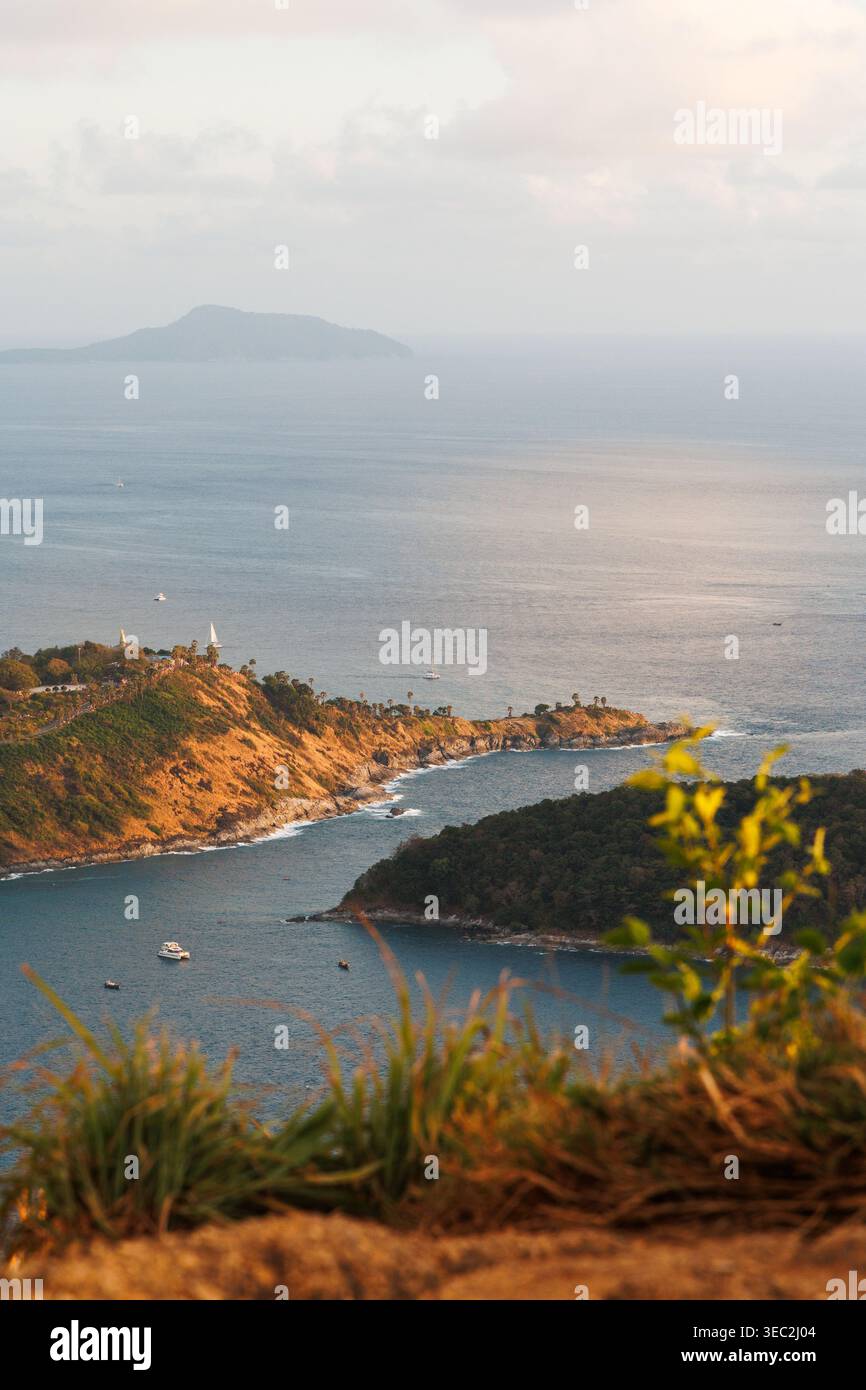 Vertikale Küstenansicht von Phuket mit verschwommener grüner Vegetation im Vordergrund, die Bucht und Landzunge einrahmt. Weiches Nachmittagslicht, orange-brauner Hügel Stockfoto