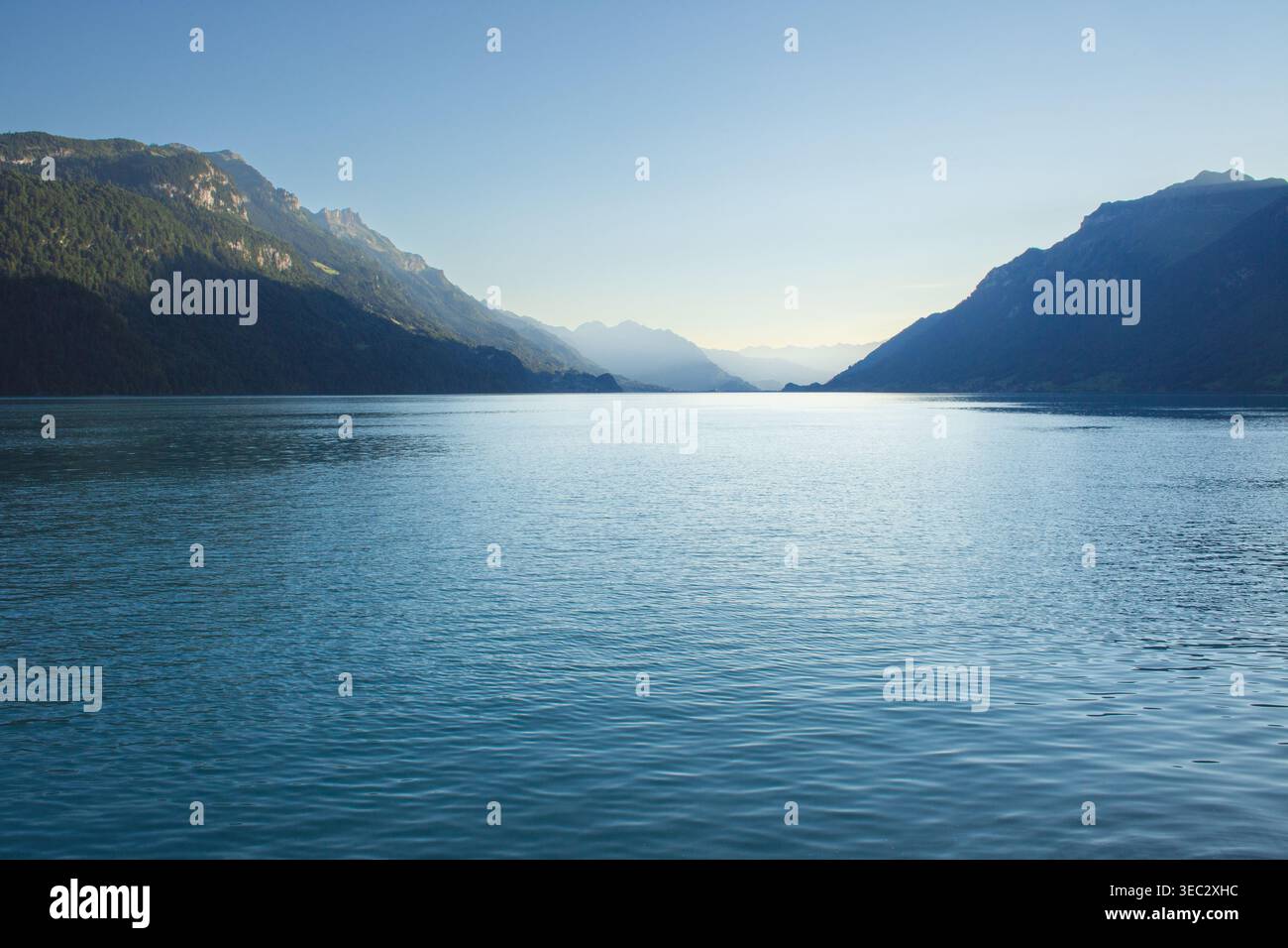 Eine weite Aufnahme eines riesigen blauen Sees auf Augenhöhe, die das sanfte Morgenlicht reflektiert, mit steilen bewaldeten Berghängen, die den Horizont unter einem klaren Kumpel umrahmen Stockfoto