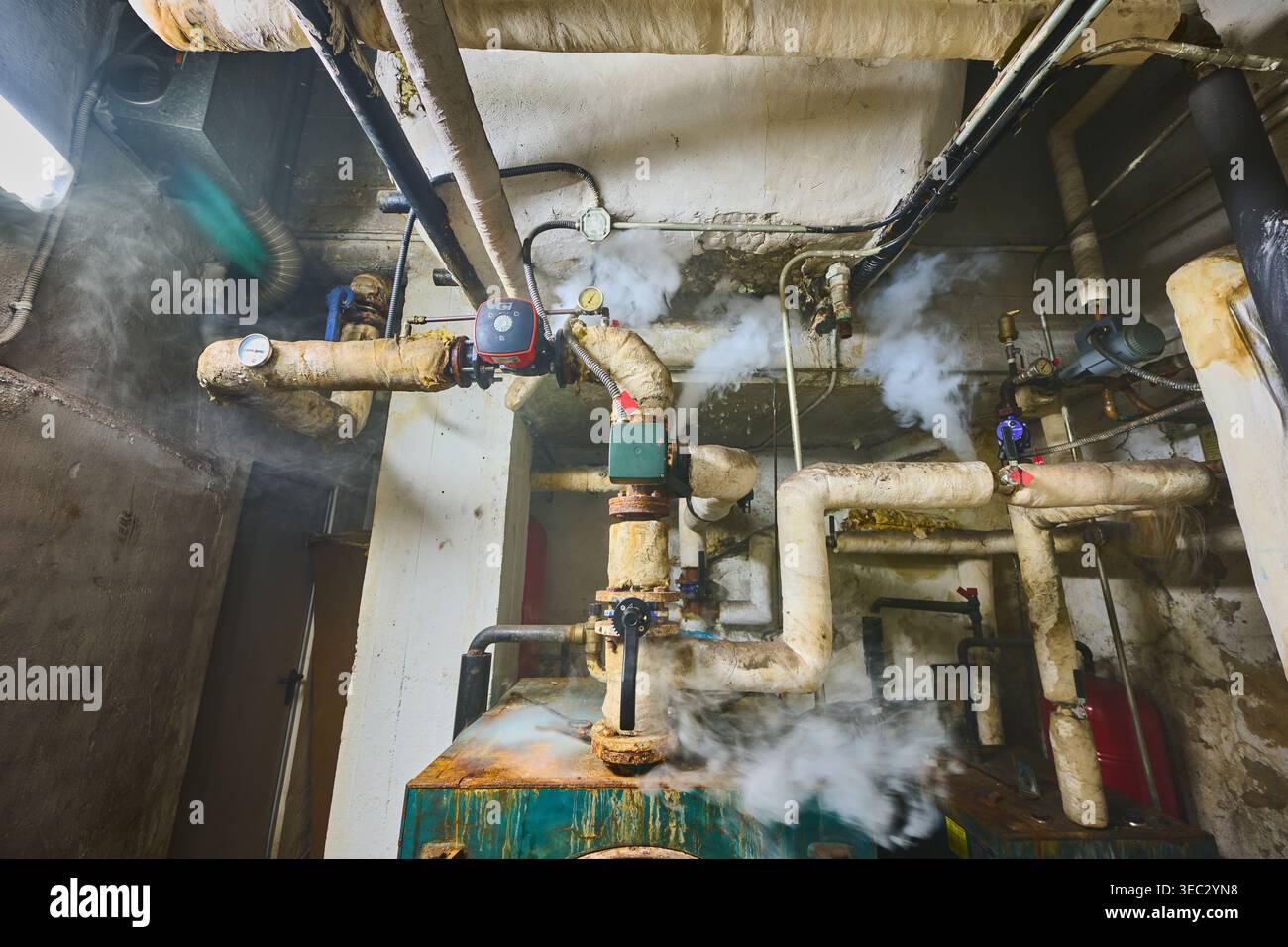 Altes, korrodiertes Kesselsystem mit isolierten Rohren, die Dampflecks aufweisen und eine gefährliche und nicht gewartete Industrieumgebung darstellen Stockfoto