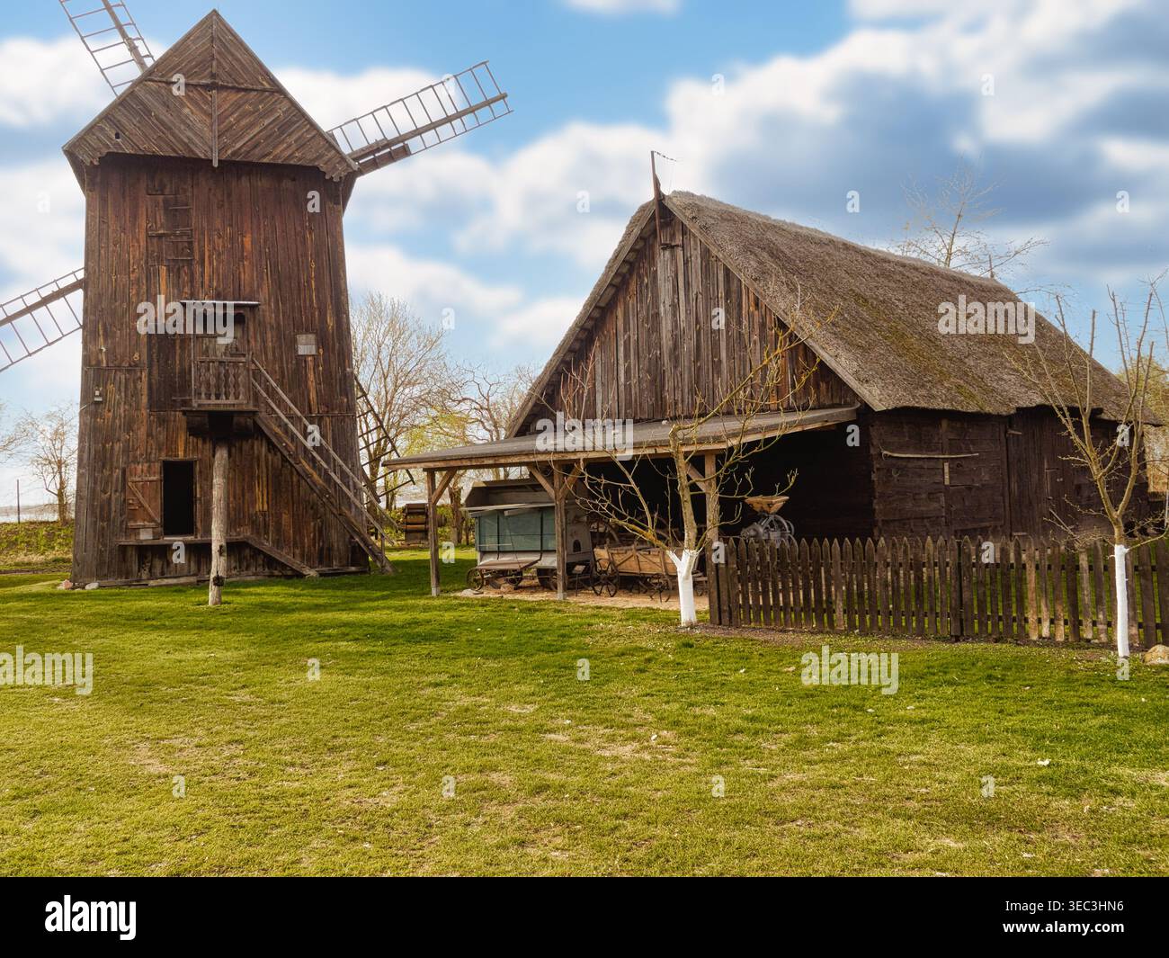 Eine traditionelle Holzwindmühle steht neben einer großen Strohdachscheune in ländlicher Umgebung. Der Hof ist von einem Holzzaun mit jungen Bäumen und umgeben Stockfoto