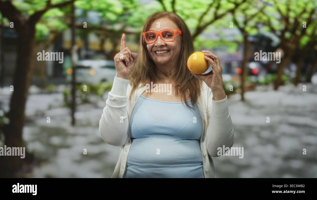 Eine Frau, die einen orangen Finger auf eine von Bäumen gesäumte Stadtstraße hält. Sie trägt eine orange Brille, ein hellblaues Tanktop und eine weiße Strickjacke Stockfoto