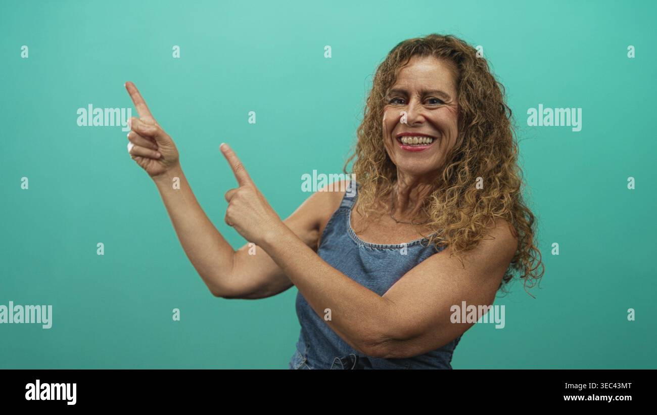 Frau mittleren Alters zeigt beide Zeigefinger im Studio nach oben, während sie breit lächelt, mit sichtbaren Zähnen und lockigem Haar; fröhliches Vertrauen. Stockfoto