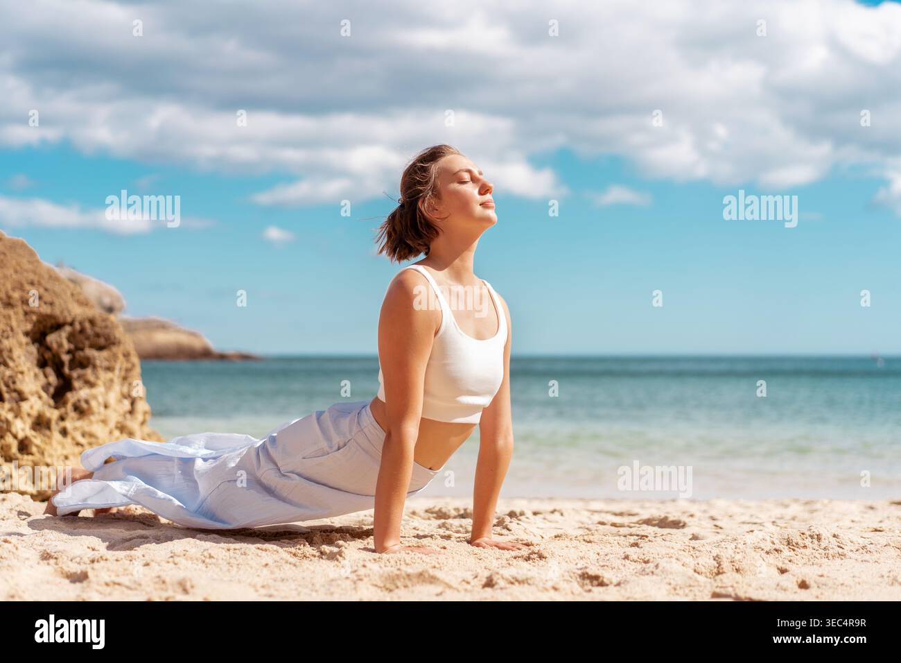 Frau in weißer Sportbekleidung, die nach oben blickt Dog Yoga Pose auf hellem Sandstrand am Meer. Sie genießt Flexibilität und Energie in ruhiger Umgebung im Freien Stockfoto