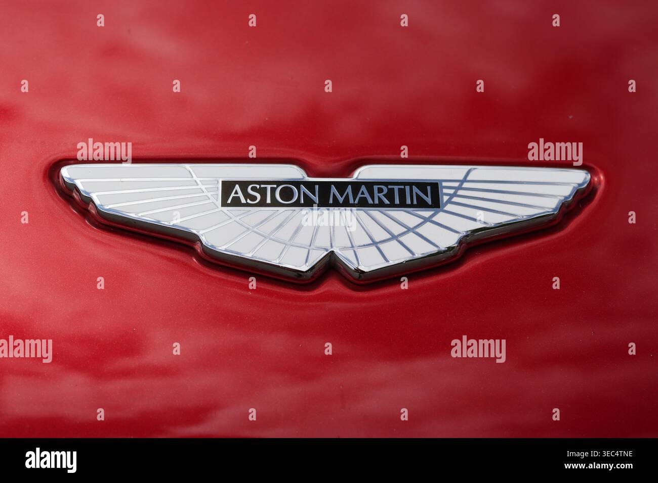 Nahaufnahme eines ikonischen Aston Martin Emblems in Emailfarbe, umgeben von einer metallischen Toro-roten Karosseriefarbe. Stockfoto