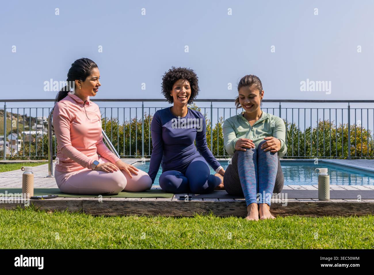 Verschiedene Freundinnen sitzen auf Yogamatten am Pool, in sportlicher Kleidung mit Wasserflaschen Stockfoto