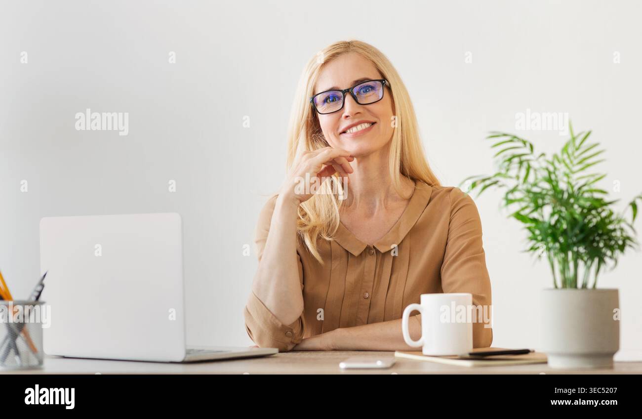 Professionelle Frau, die zu Hause arbeitet, lächelt, während sie an einem aufgeräumten Schreibtisch mit Laptop sitzt Stockfoto