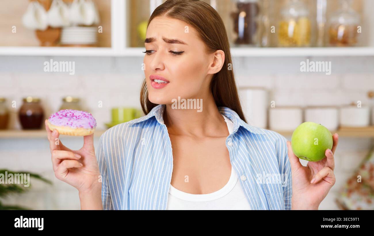 Frau hält Apfel und Donut, während sie zwischen gesunden und süßen Optionen in der Küche entscheidet Stockfoto
