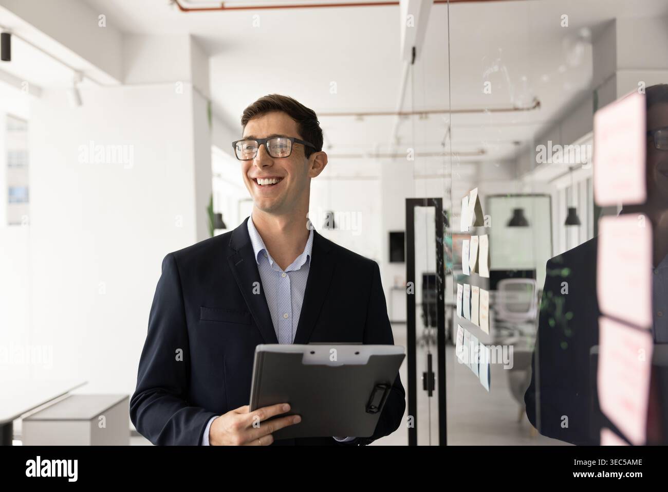 Fröhlicher Geschäftsmann in Anzug steht in einem modernen Büro mit Klemmbrett Stockfoto