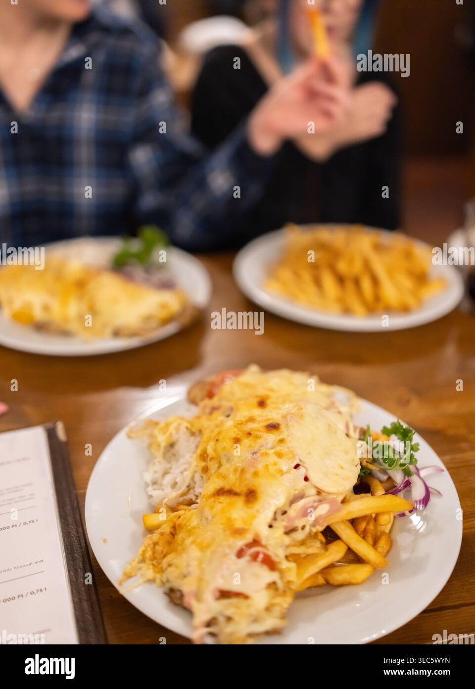 Herzhafte Mahlzeit mit goldenen Pommes frites. Käsegericht mit frischen Kräutern. Die Person im karierten Hemd hält eine Gabel. Der Holztisch bietet Platz für Teller und Gerichte. Foo Stockfoto
