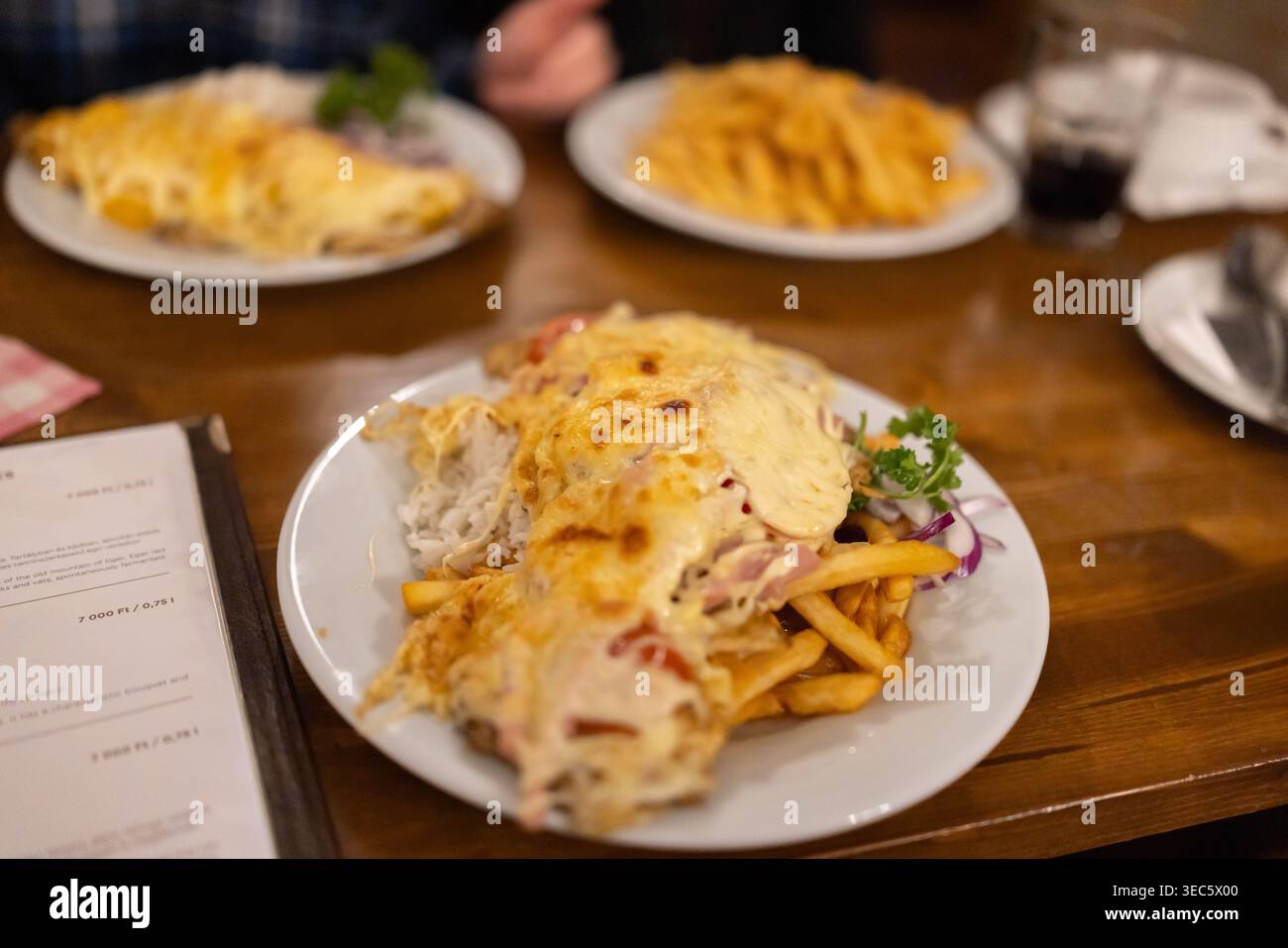 Goldenes Omelette mit Käse über knusprigen Pommes frites. Frische Petersilie-Garnitur verleiht Farbe. Teller sitzen auf einem warmen Holztisch. Die Dinnerszene erinnert an Komfort und Unterhaltung Stockfoto
