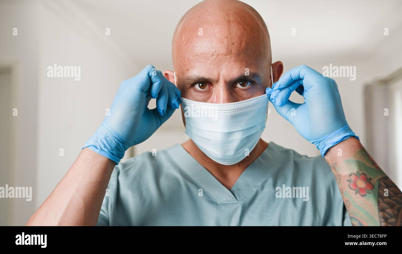Healthcare Worker, der chirurgische Maske und Handschuhe in klinischer Umgebung mit tätowiertem Arm aufsetzt Stockfoto