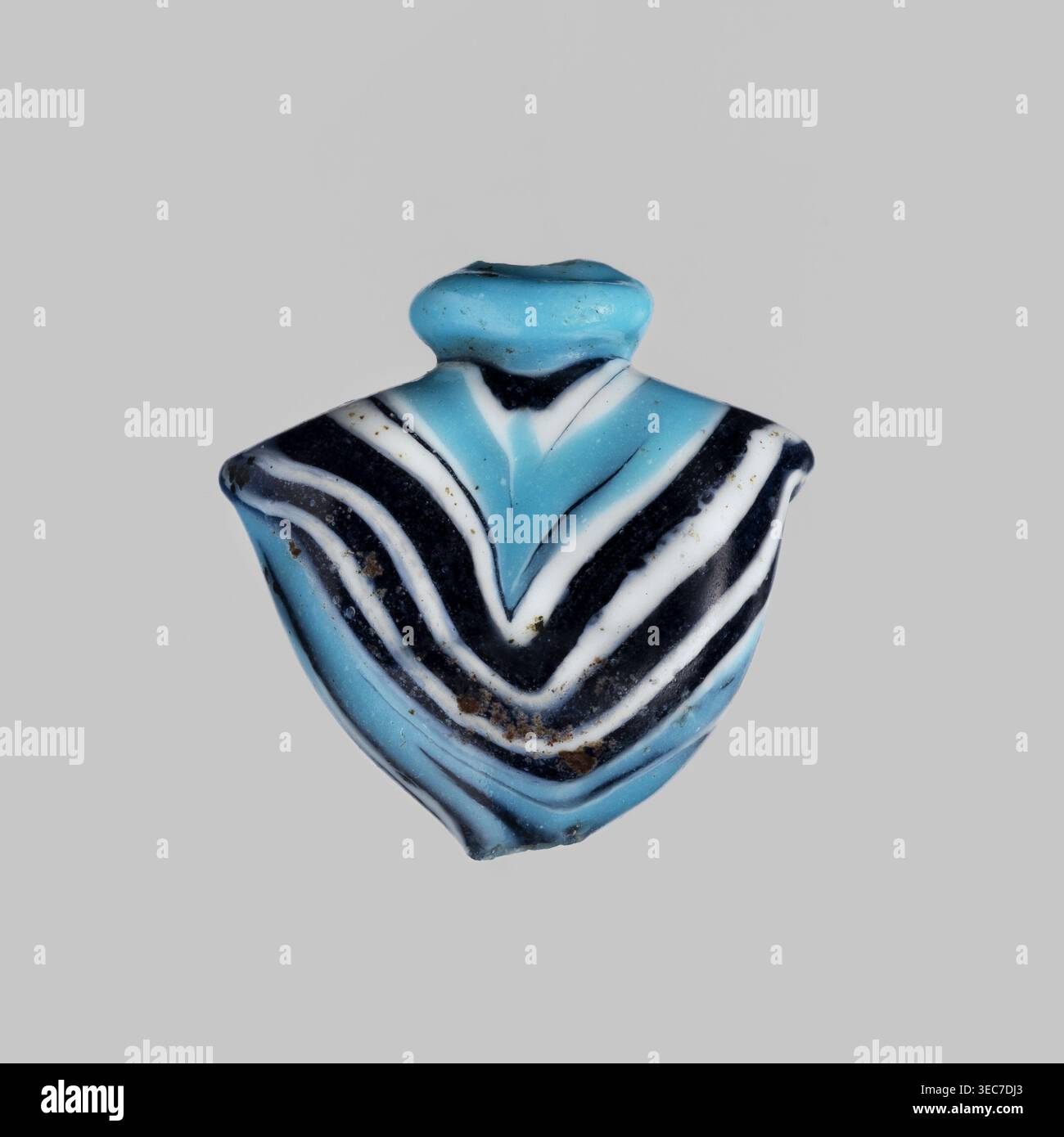 Amulett in Form eines Herzens aus blauem Glas, verziert mit parallelen Linien, die vorne und hinten einen Zickzack bilden, in Marineblau und weiß. Das Amulett ist senkrecht durchbohrt, das Loch oben mit einem blauen Ring umrahmt, an den Seiten verbleibende Griffe. 1550–1295 v. Chr.; Neues Königreich, 18. Dynastie, Ägypten. Amulett Heart/IB, Höhe 1,9 cm, Breite 1,9 cm., Ein kleines Objekt mit einer schildähnlichen Form, zentriert auf einem glatten hellgrauen Hintergrund, das geschwungene Bänder in hellem Türkis, tiefem Schwarz und weiß zeigt, die ein Chevron-Muster bilden, mit einem abgerundeten türkisfarbenen oberen Abschnitt; die Oberfläche zeigt Stockfoto