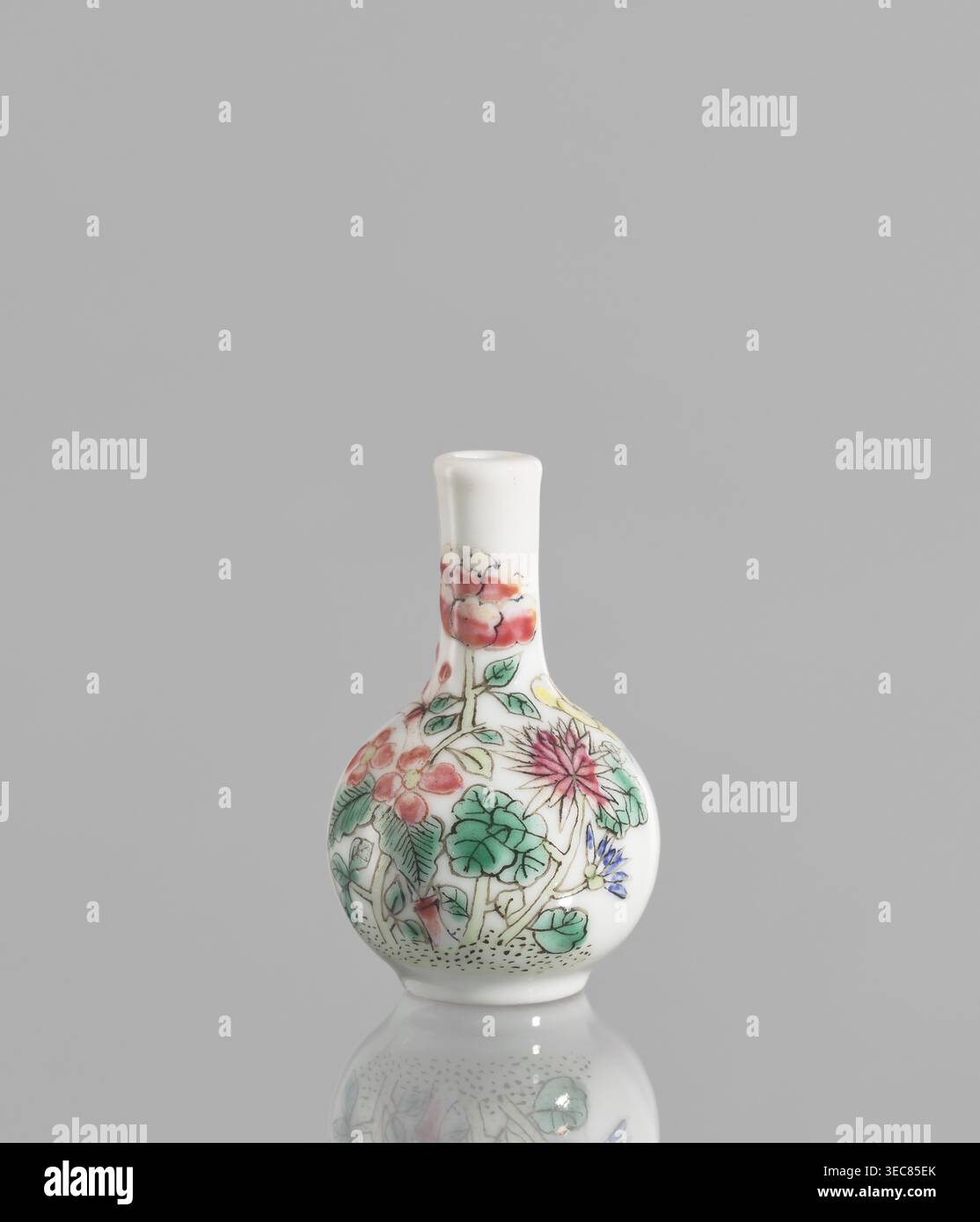 Flasche, Wiederbesitz Krakau, Keramik; eine sehr kleine eiförmige Flasche mit langem, dünnem, geradem Hals. Weiße Glasur mit Emailfarben und schwarzer Konturüberglasung. Nicht Signiert. 19./20. Jahrhundert, China. Gefäß. Höhe 5,7 cm; Durchmesser 3,5 cm., Eine kleine Porzellanflasche mit einem abgerundeten Körper und einem schmalen Hals sitzt auf einem glatten grauen Hintergrund, deren weiße Oberfläche mit einem bemalten Blumenmuster verziert ist, das eine Gruppe von Blüten und Blättern in den Farben Rosa, Rot, Grün, Blau, Gelb zeigt, schwarze Umrisse und die Flasche und ihre bemalten Motive spiegeln sich sanft auf der glänzenden Oberfläche wider Stockfoto