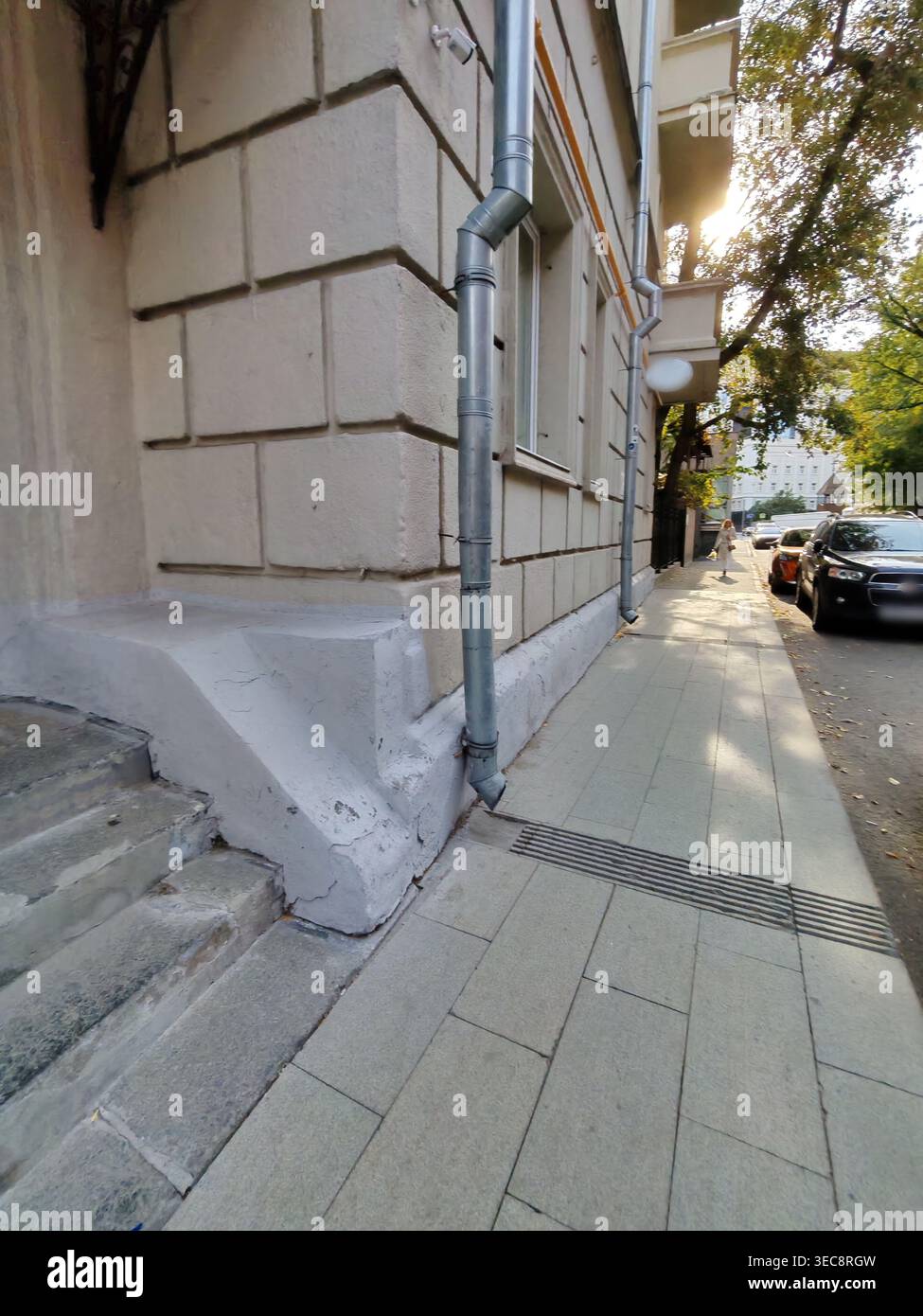 Eine Nahaufnahme einer verwitterten Gebäudeecke mit sichtbaren Abflussrohren, gerissenen Steintreppen und einem gekachelten Bürgersteig. Auf dem Foto werden städtische Details und die erfasst Stockfoto