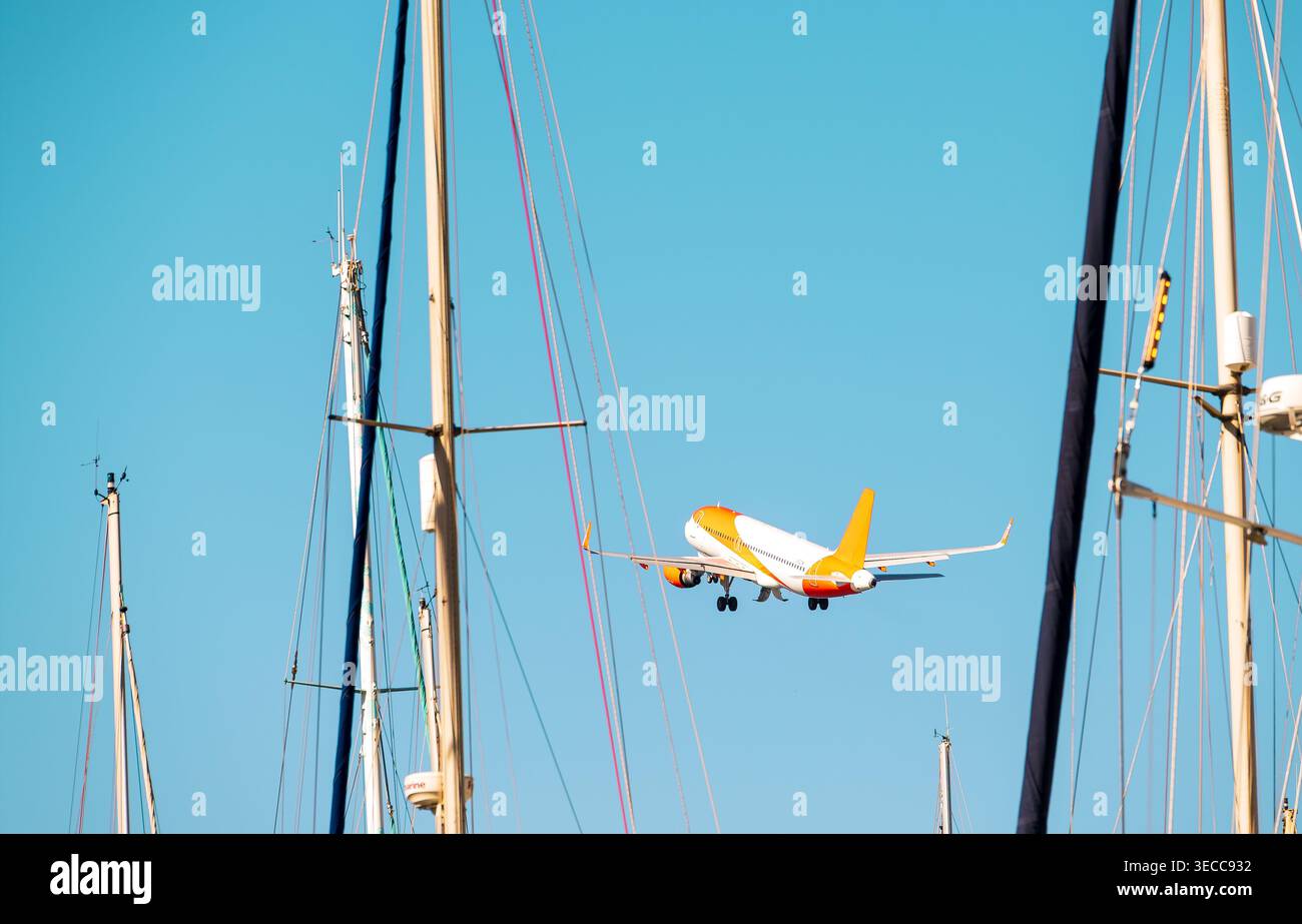 Ein kommerzielles Flugzeug mit orangefarbener und weißer Lackierung fliegt tief, eingerahmt von Masten und Takelage mehrerer Segelboote. Der Himmel ist klar und blau, Sug Stockfoto