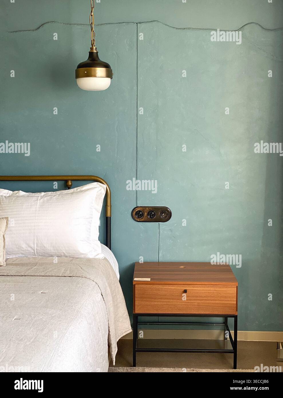 Minimales Innendetail des Schlafzimmers mit Bett, hölzernem Nachttisch und Pendelleuchte an strukturierter blauer Wand - Smartphone-aufgenommenes Stockfoto