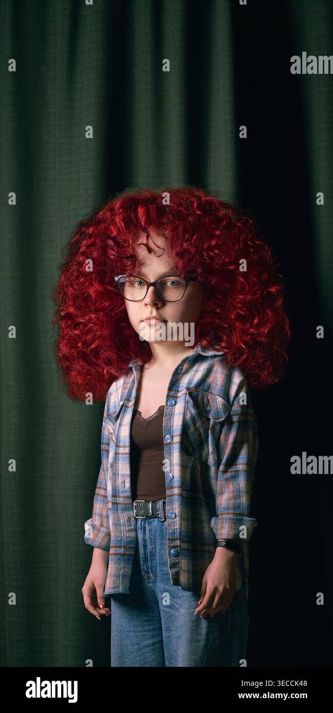Mädchen mit übergroßen roten Locken und Brille steht vor dunkelgrünem Vorhang. Stockfoto