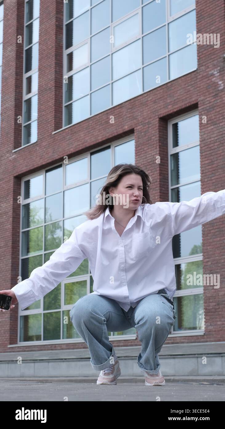 Junge Frau in der Hocke mit gestreckten Armen und konzentrierter Haltung im städtischen Umfeld. Die dynamische Szene zeigt emotionale Spannung und Kontrolle Stockfoto