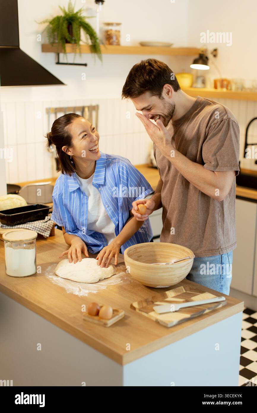In einer hellen Küche lachen eine Koreanerin und ein kaukasischer Mann beim Backen. Jede freudige Interaktion zeigt ihre Verbindung durch Essen. Stockfoto