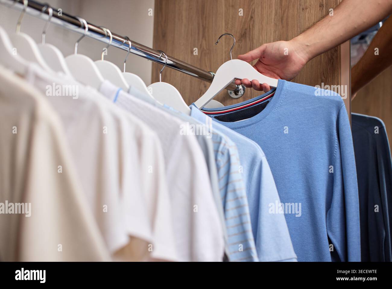 Wählen Sie von Hand ein hellblaues T-Shirt mit einem weißen Kleiderbügel aus einer Auswahl an Kleidungsstücken auf einem Kleiderständer in einem Holzschrank aus Stockfoto