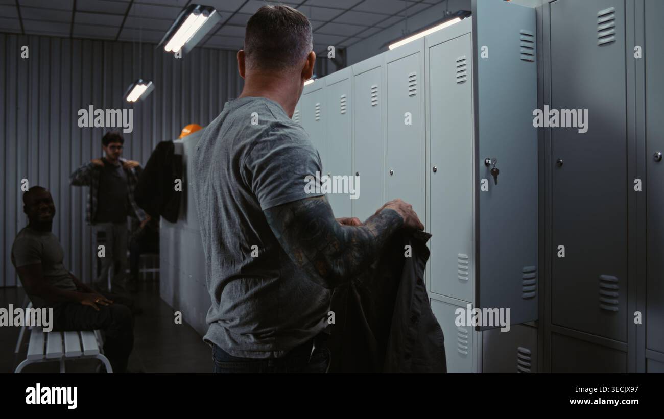 Drei verschiedene Industriearbeiter in High Visibility Gear Friendly interagieren, lachen und teilen Witze, während sie sich für ihre Schicht in der modernen Fabrik kleiden. Unternehmenskultur und Teamgeist. Stockfoto