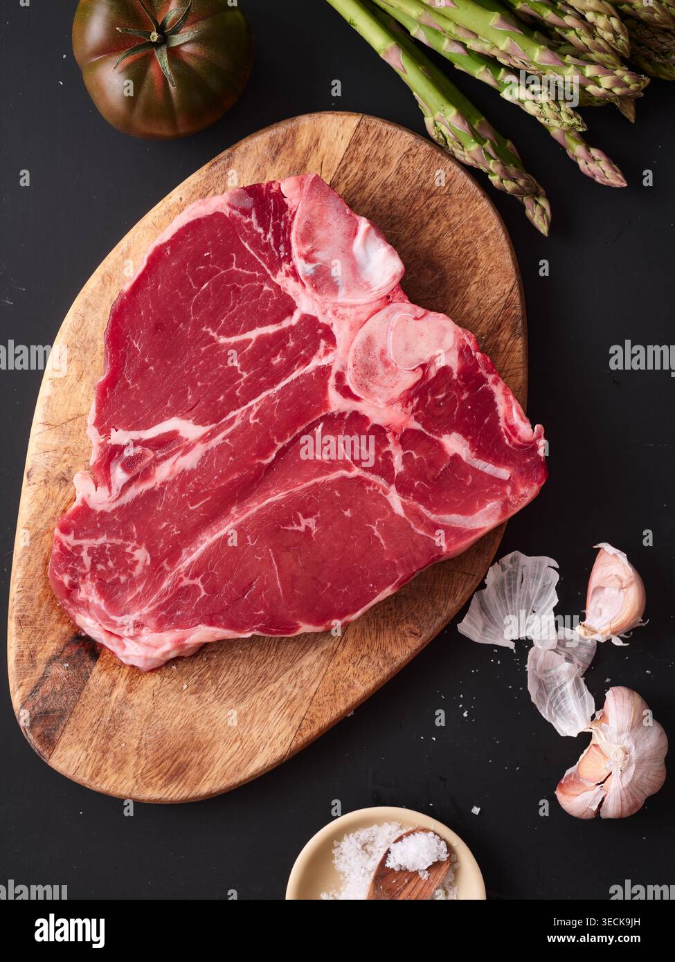 Ein rotes T-Bone-Steak sitzt auf einem hölzernen Schneidebrett. Daneben sind Knoblauchzehen und frisches Gemüse im Hintergrund. Diese Einstellung ist für eine Mahlzeit p Stockfoto