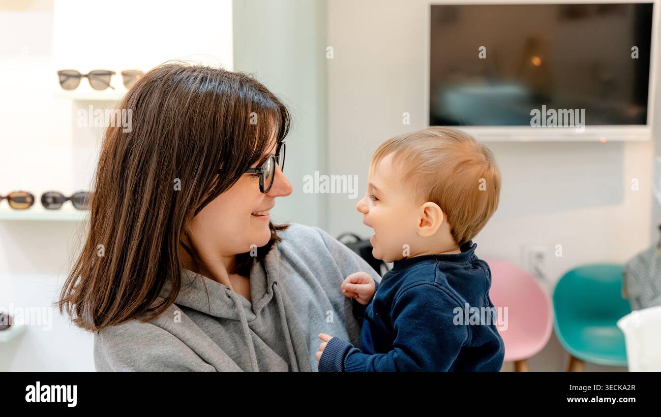 Eine fröhliche Mutter mit Brille teilt einen herzlichen Moment mit ihrem lächelnden Baby in einem optischen Zentrum, das Wärme und Kundenzufriedenheit in einem widerspiegelt Stockfoto