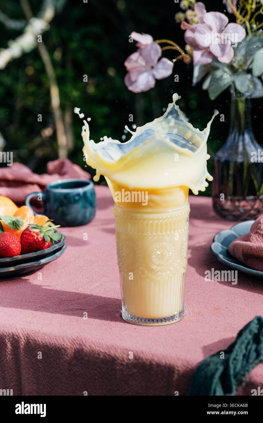 Orangen-, Ananas- und Aprikosensaft, der in einem hohen Glas auf einem Frühstückstisch mit frischem Obst, Blumen und sanftem Morgenlicht sprudelt und eine Vinnie schafft Stockfoto