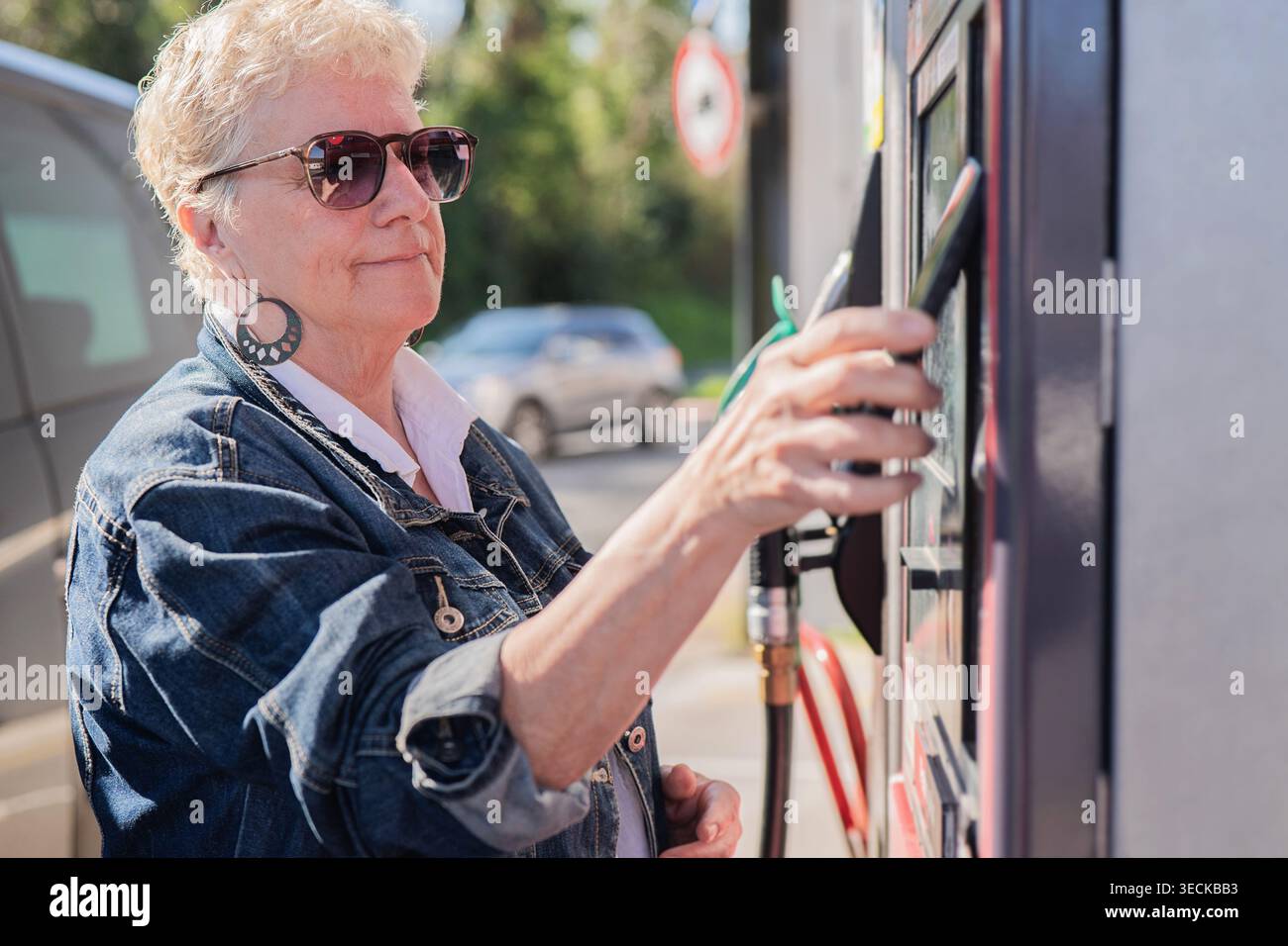 Eine ältere Frau mit Sonnenbrille und Jeansjacke benutzt eine Tankstelle und bezahlt mit ihrem Handy. Die Szene erfasst einen Stockfoto