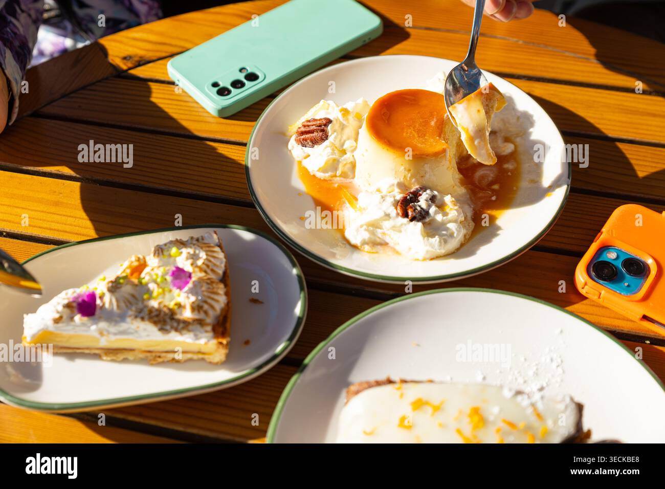 Sonnendurchfluteter Terrassentisch mit cremigem Flan, Karamell, Schlagsahne und Nüssen sowie einem Stück Kuchen. Smartphones ruhen in der Nähe, was auf eine zwanglose Art hindeutet Stockfoto